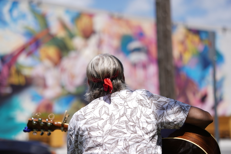 A man plays guitar in front of the Russell Street Parkette Mural.