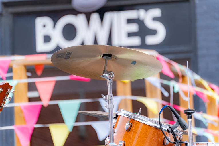 A shot of a drum kit on stage with the Bowie's Music Club's sign in the background.